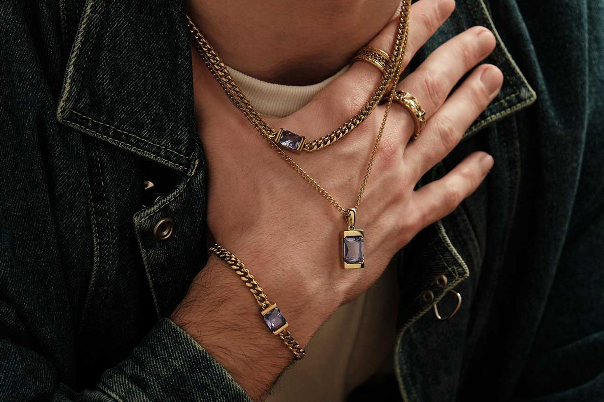 Man in a denim jacket wearing a gold Cuban chain layered over a pendant chain, two stacked gold rings and a gold Cuban bracelet